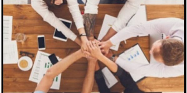 Team members join hands in a show of unity over a workspace with documents and devices.