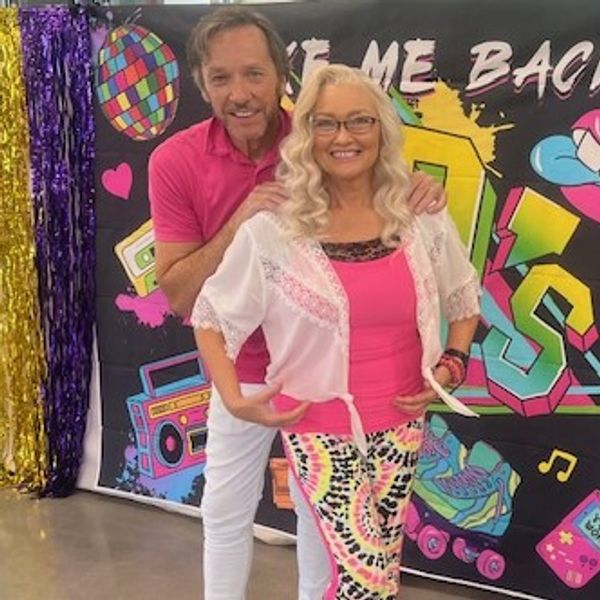 A smiling couple posing in colorful 80s-themed outfits with a vibrant backdrop.