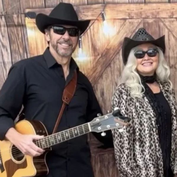Two people in cowboy hats, one playing guitar, standing in front of wooden backdrop.