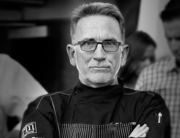 Serious chef in black uniform posing with arms crossed in a professional setting.