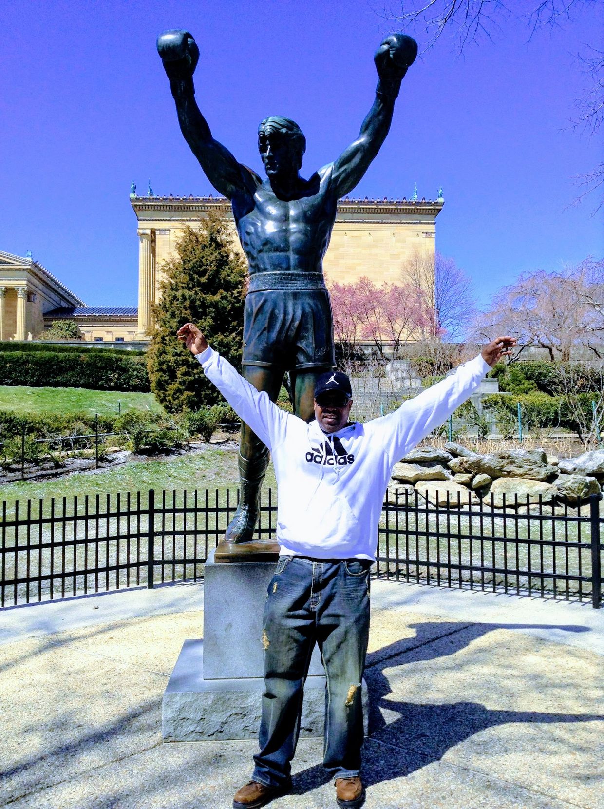 Man posing with a bronze Rocky Balboa statue outdoors.