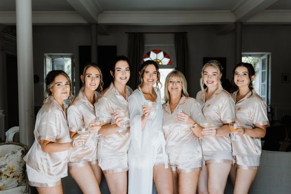 Bride and bridesmaids in matching silk robes holding drinks indoors.