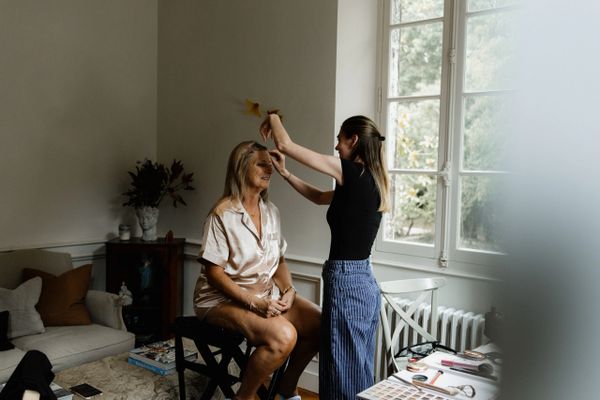 A woman applies makeup on another woman sitting by a window in a cozy room.