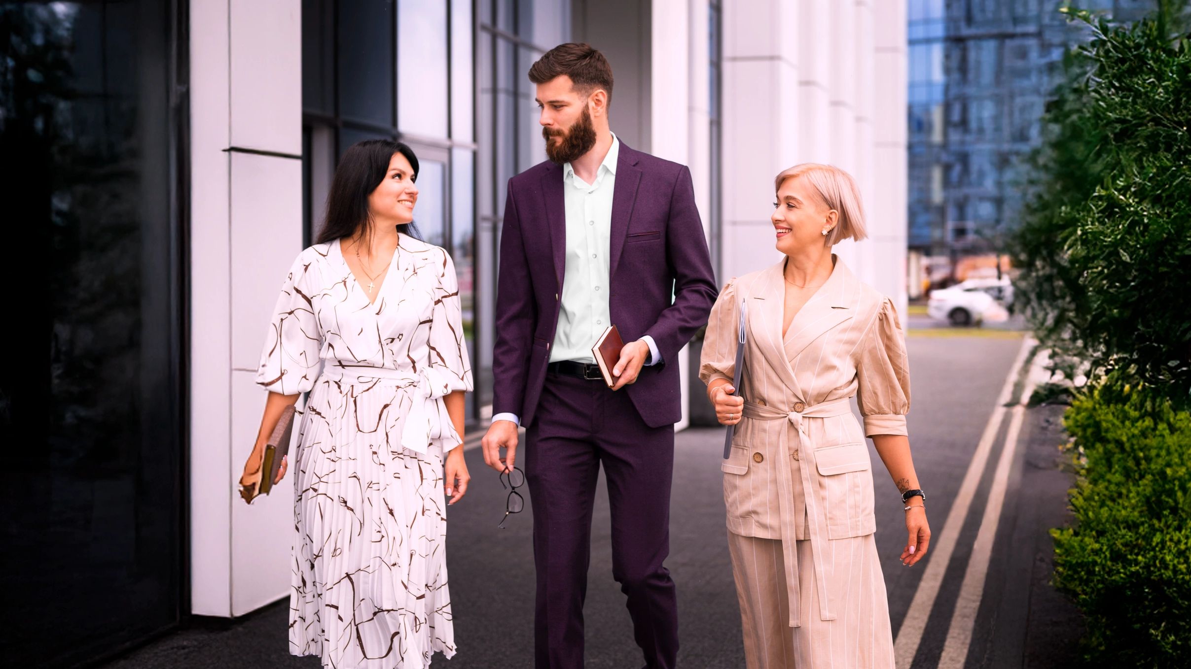 Three adults walk by an office building, talking. They appear confident, with plants & glass nearby.