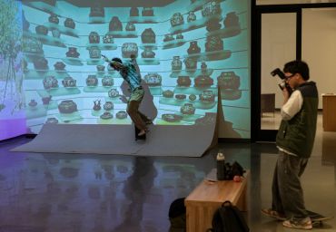 Person posing on a ramp while another takes photos in a gallery-like room.