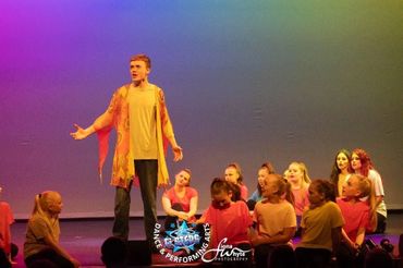 A young performer passionately sings on stage with a group of seated dancers.