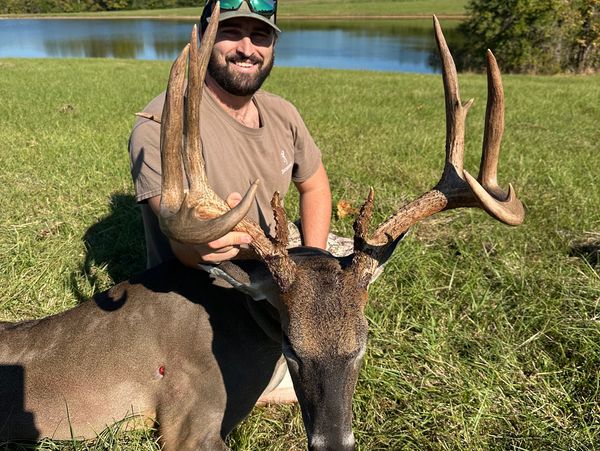 Hunter proudly poses with a large buck beside a scenic lake on a sunny day.