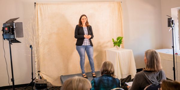 Woman giving a presentation on a small stage in front of seated audience.