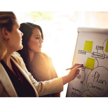 Two women discussing a plan on a flip chart in a bright office.