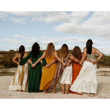 Six women standing arm in arm in a desert setting, wearing colorful dresses.