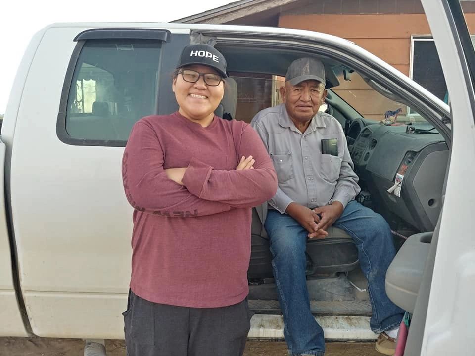 Two men posing with a white truck, one standing smiling with a 'HOPE' cap, the other seated inside.