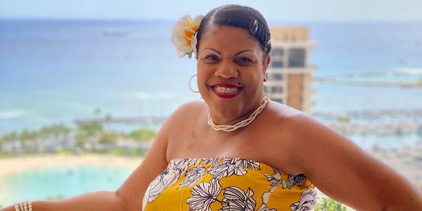 Woman in a yellow floral dress smiling by a balcony with ocean view.