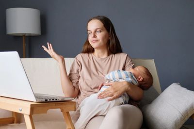 A woman holding a baby while looking at a laptop with a thoughtful expression.