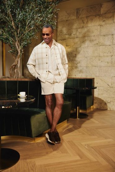 Man in casual beige outfit with sunglasses standing indoors near green velvet seating.