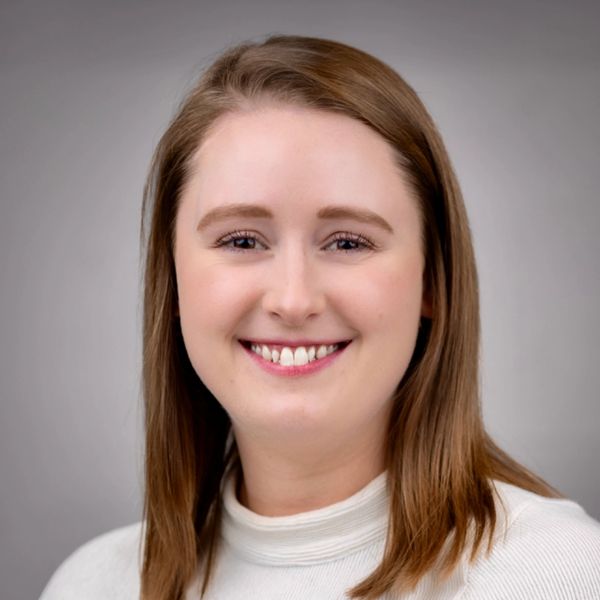 Smiling woman with light brown hair in a white top against a gray background.