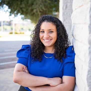 Woman with curly hair in a blue blouse smiling confidently by a stone wall.