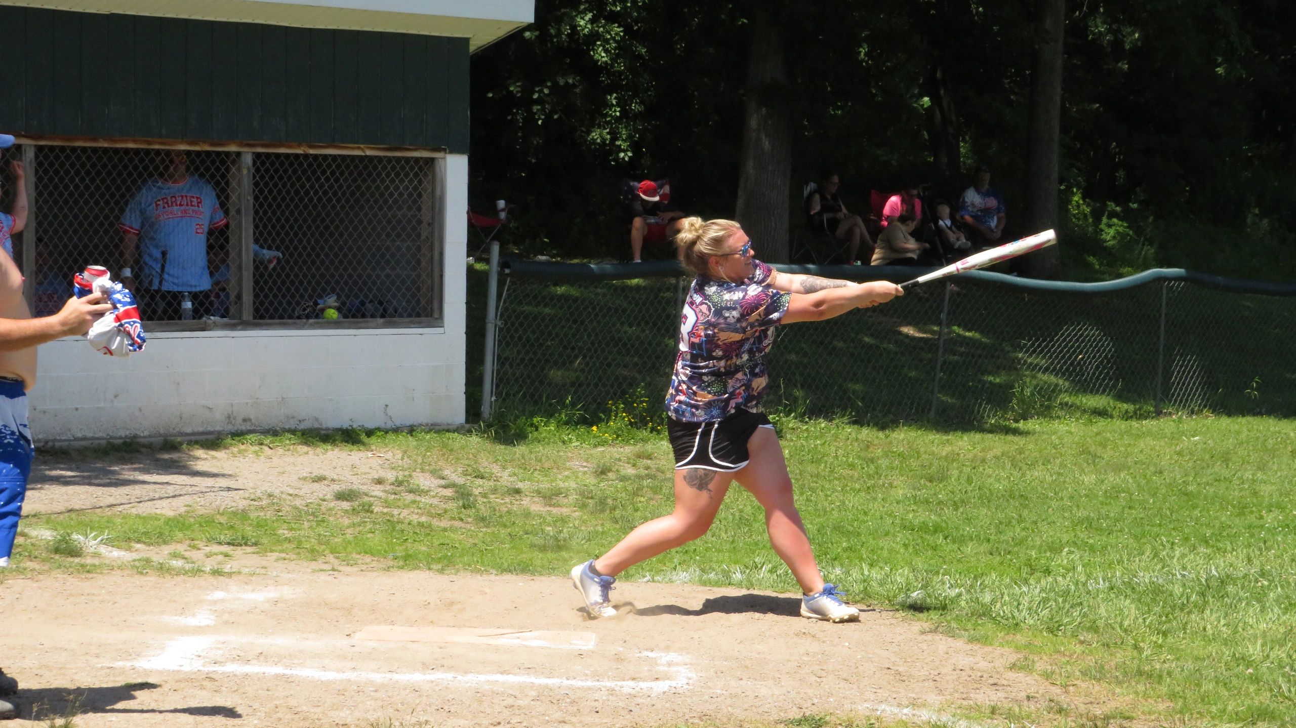 Woman in floral shirt swinging a bat on a sunny softball field.