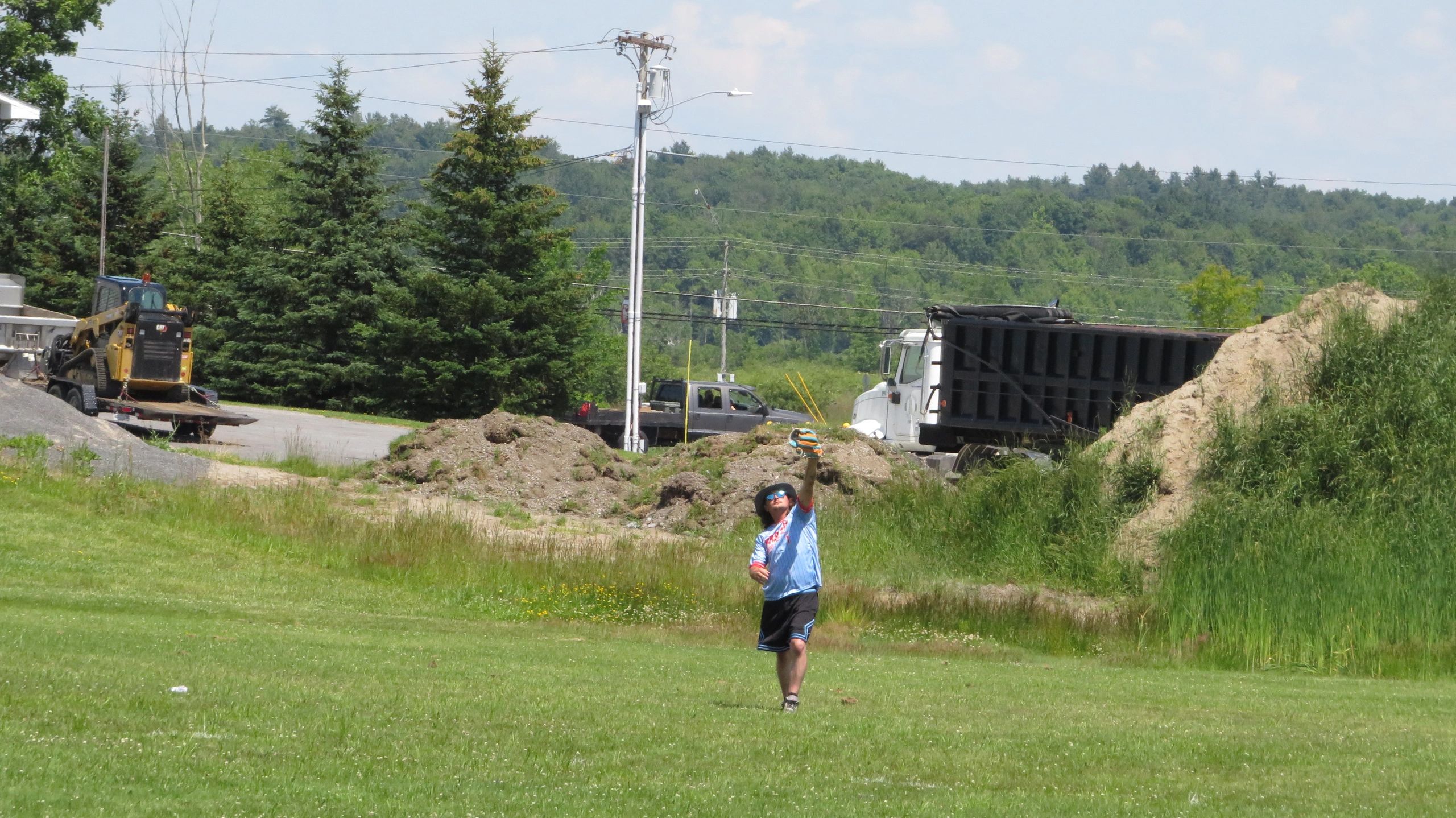 A person catches a ball in a grassy area with trucks and trees in the background.