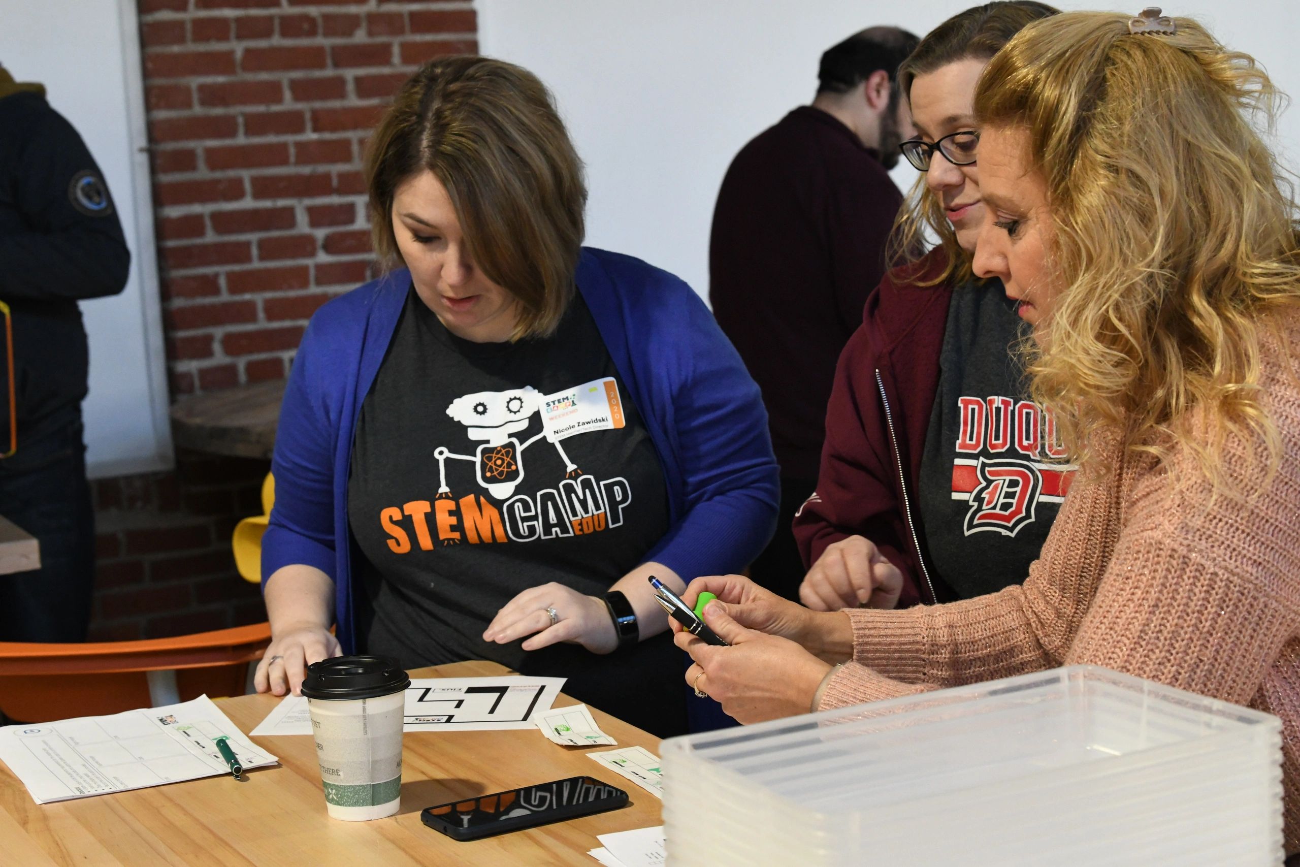 Three women engaged in a STEM camp activity at a table with papers and tools.