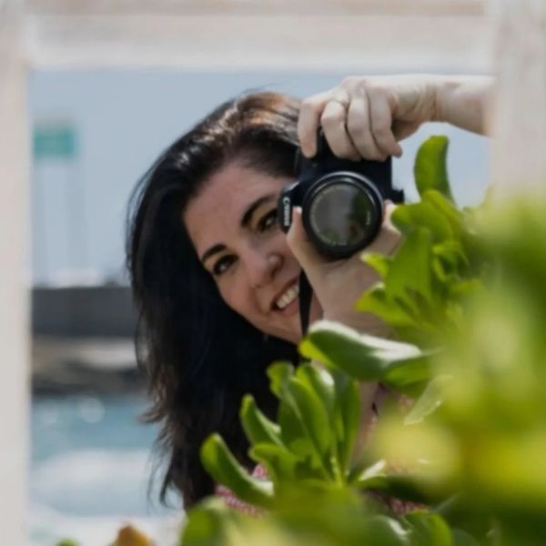 Woman smiling while holding a camera behind green leaves.