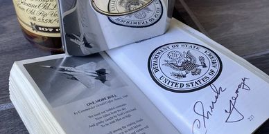 Open book showing a signed page with the US Department of State seal and a photo of a jet.