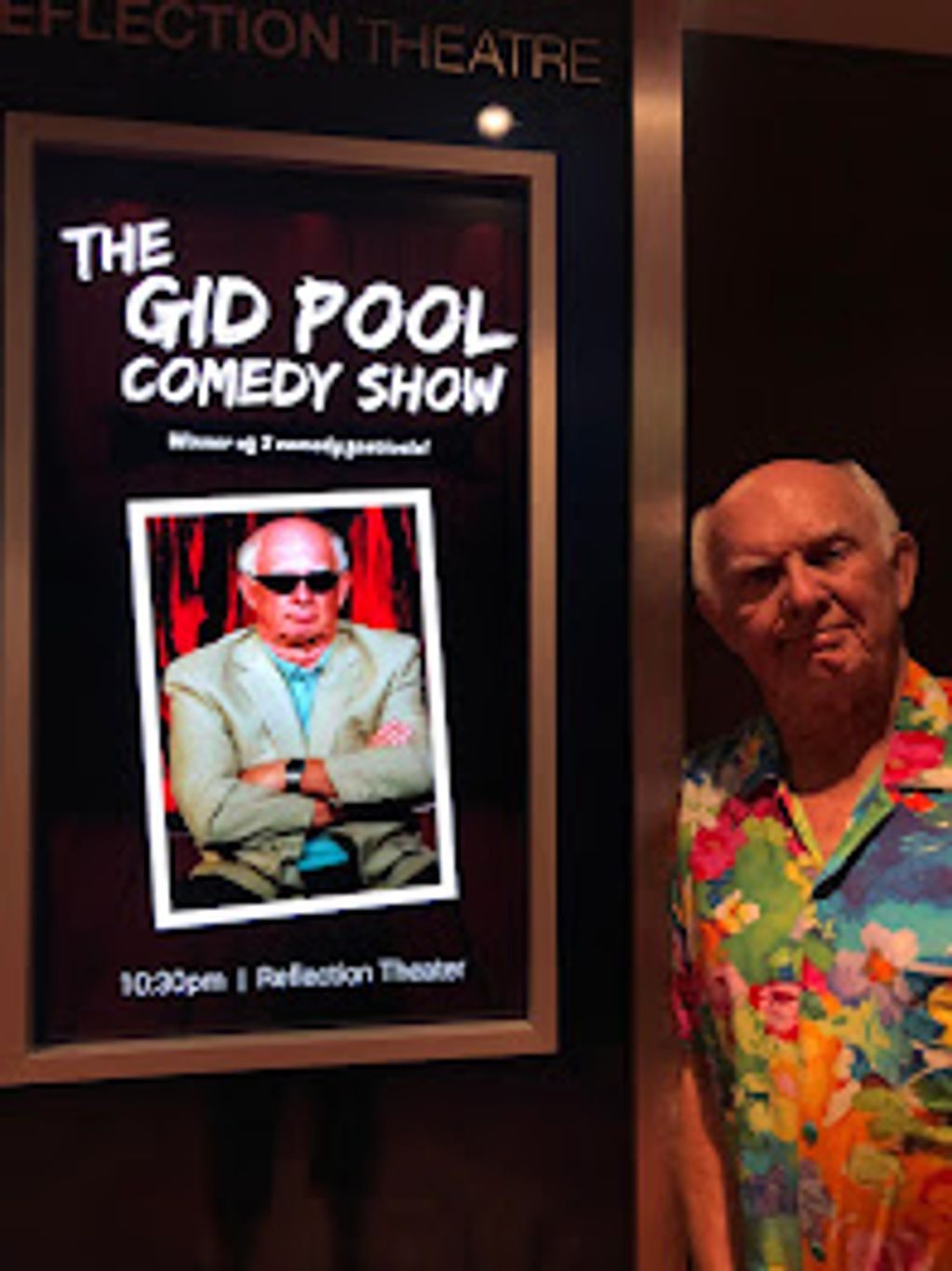 A man in a colorful shirt stands next to a comedy show poster.