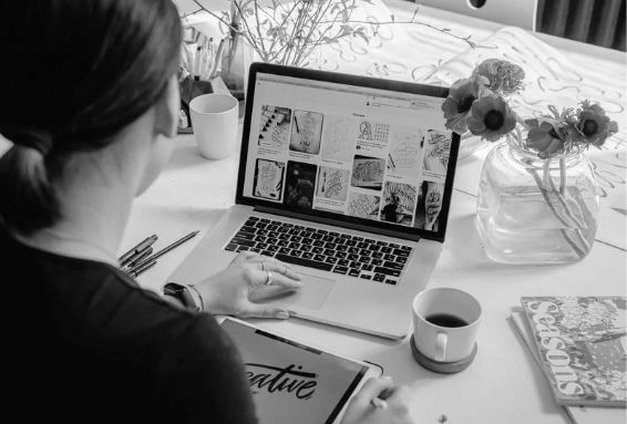 A person using a laptop and tablet, surrounded by flowers and coffee.