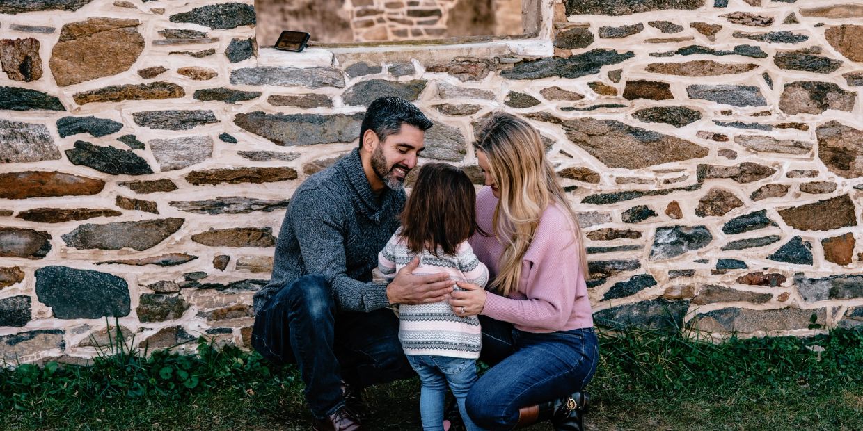 A family lovingly embraces their young child outdoors by a stone wall.