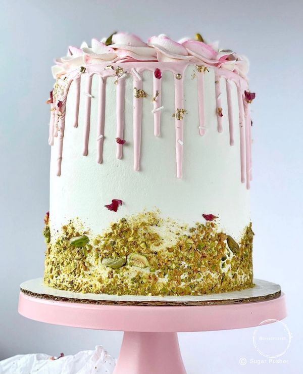 Tall cake with smooth white buttercream, pink chocolate drip, edible rose petals, crushed pistachios