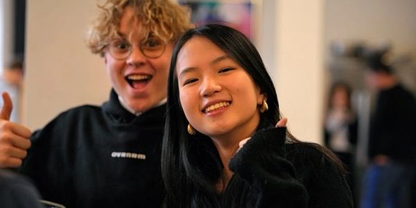 Two friends smiling, one giving a thumbs-up gesture indoors.