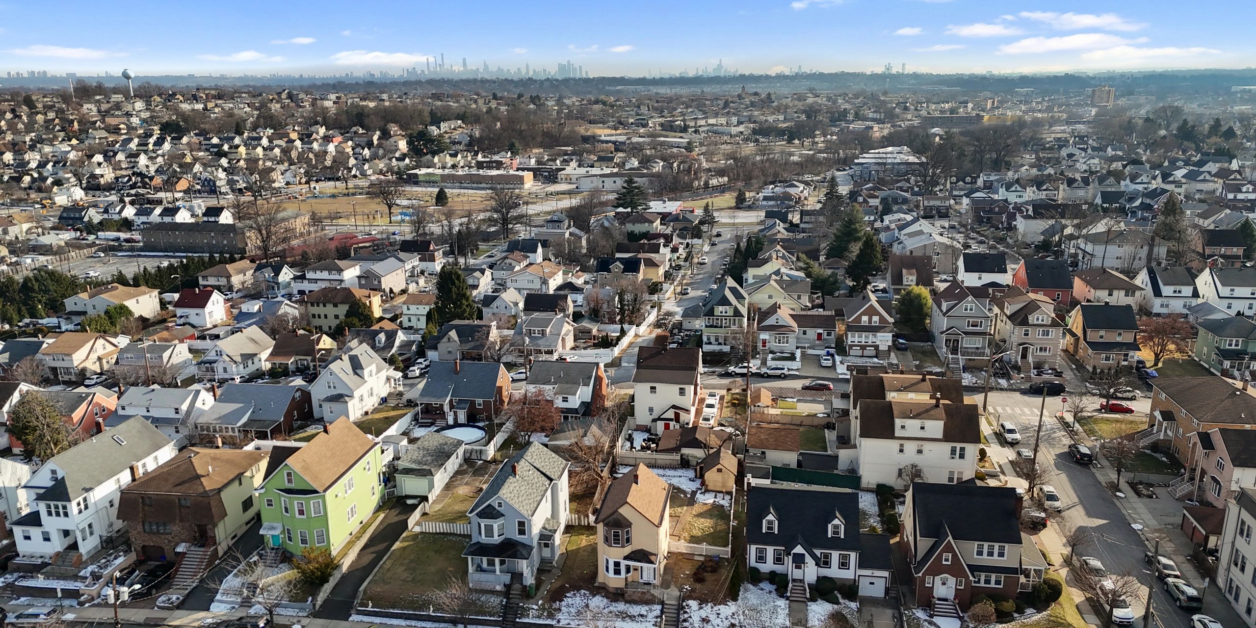 Suburban neighborhood with many houses and a city skyline in the distance under a clear sky.