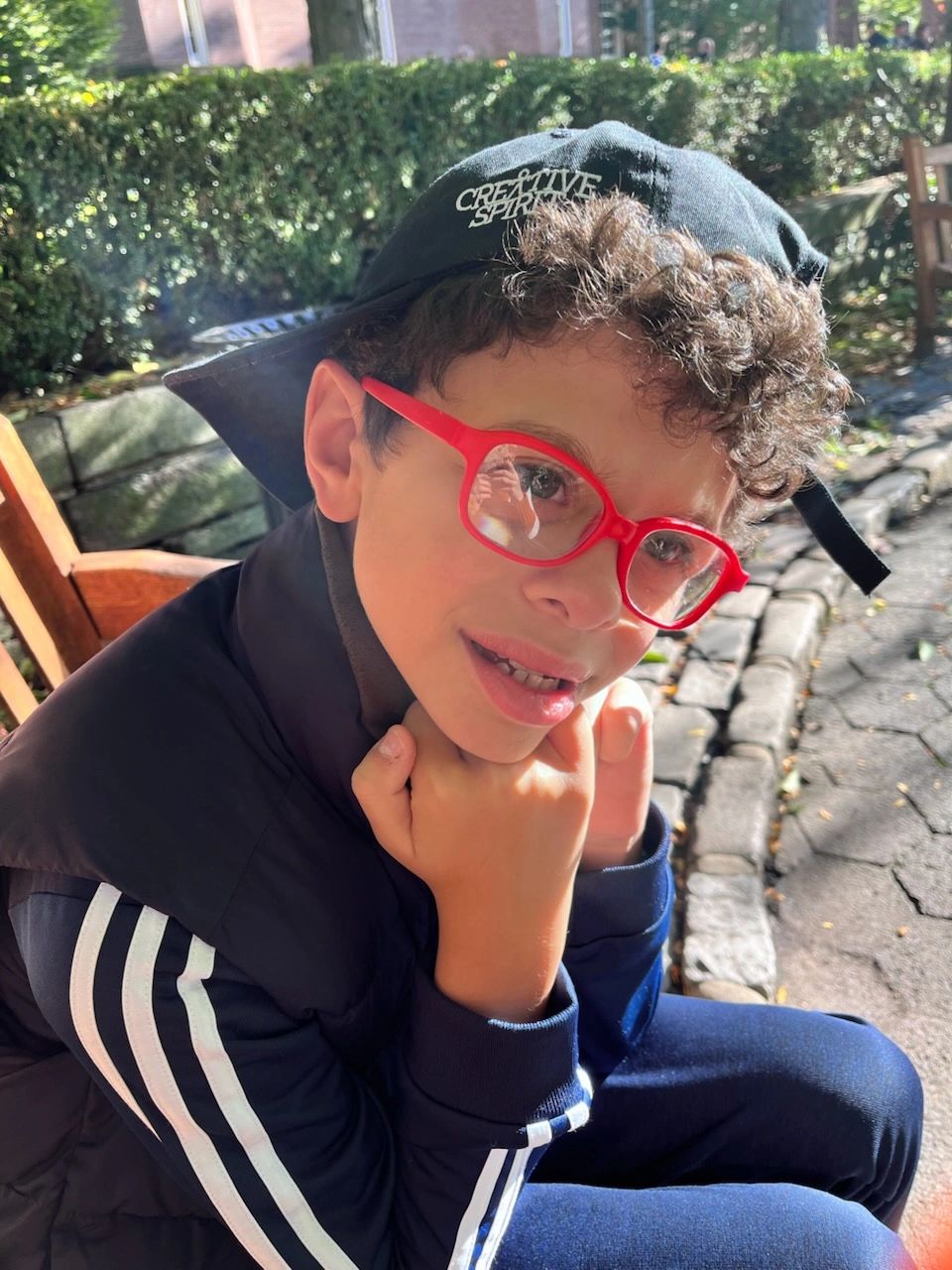 Boy with red glasses and a black cap sitting outdoors.