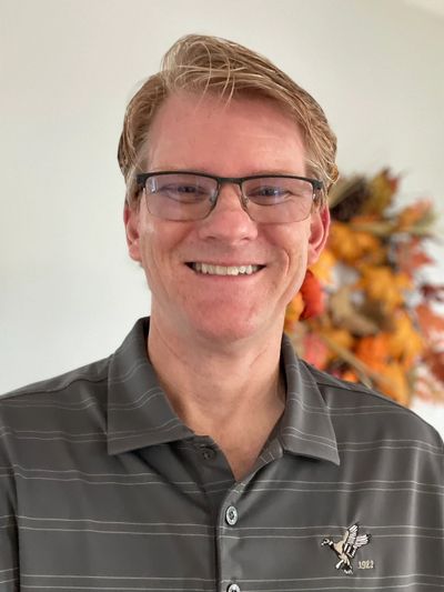 Smiling man wearing glasses and a striped polo shirt with autumn decorations in the background.