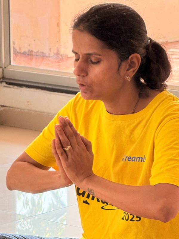 A woman in a yellow shirt with hands pressed together in a prayer pose, sitting with eyes closed.