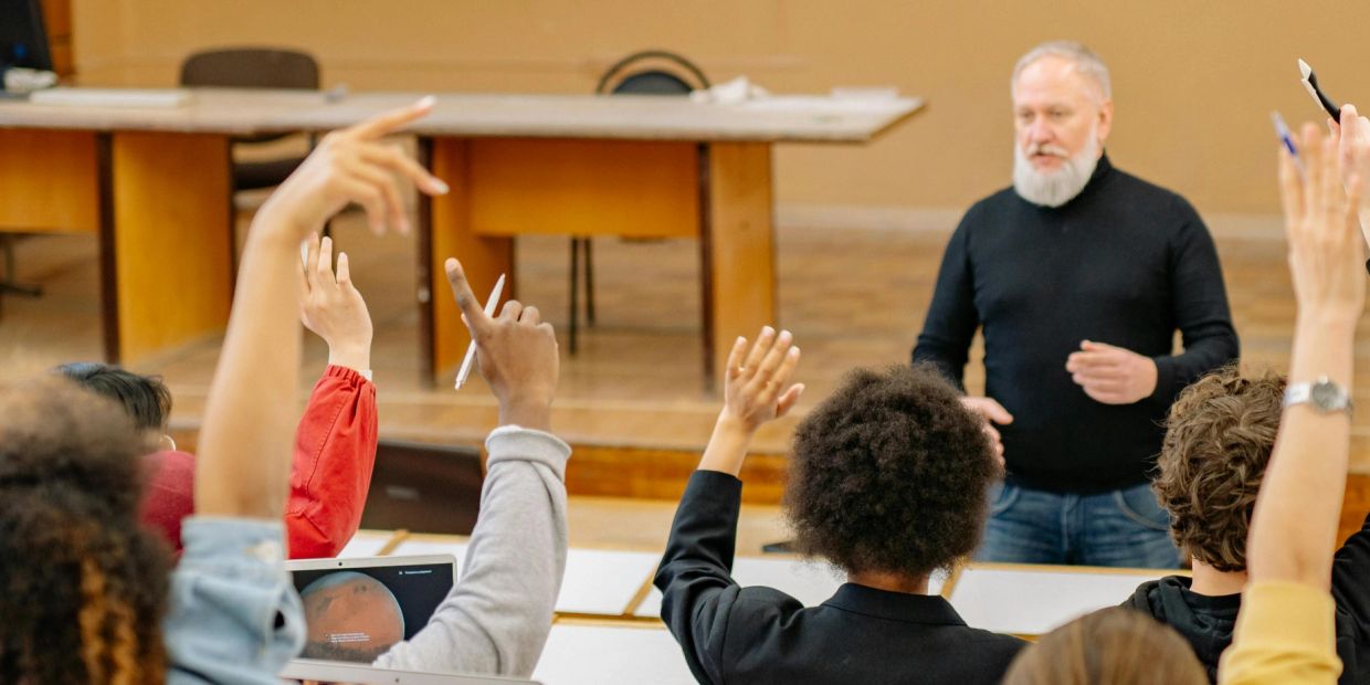 Students raising hands in a classroom with a male teacher.