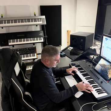 Man playing a keyboard in a music studio surrounded by multiple keyboards and a computer.