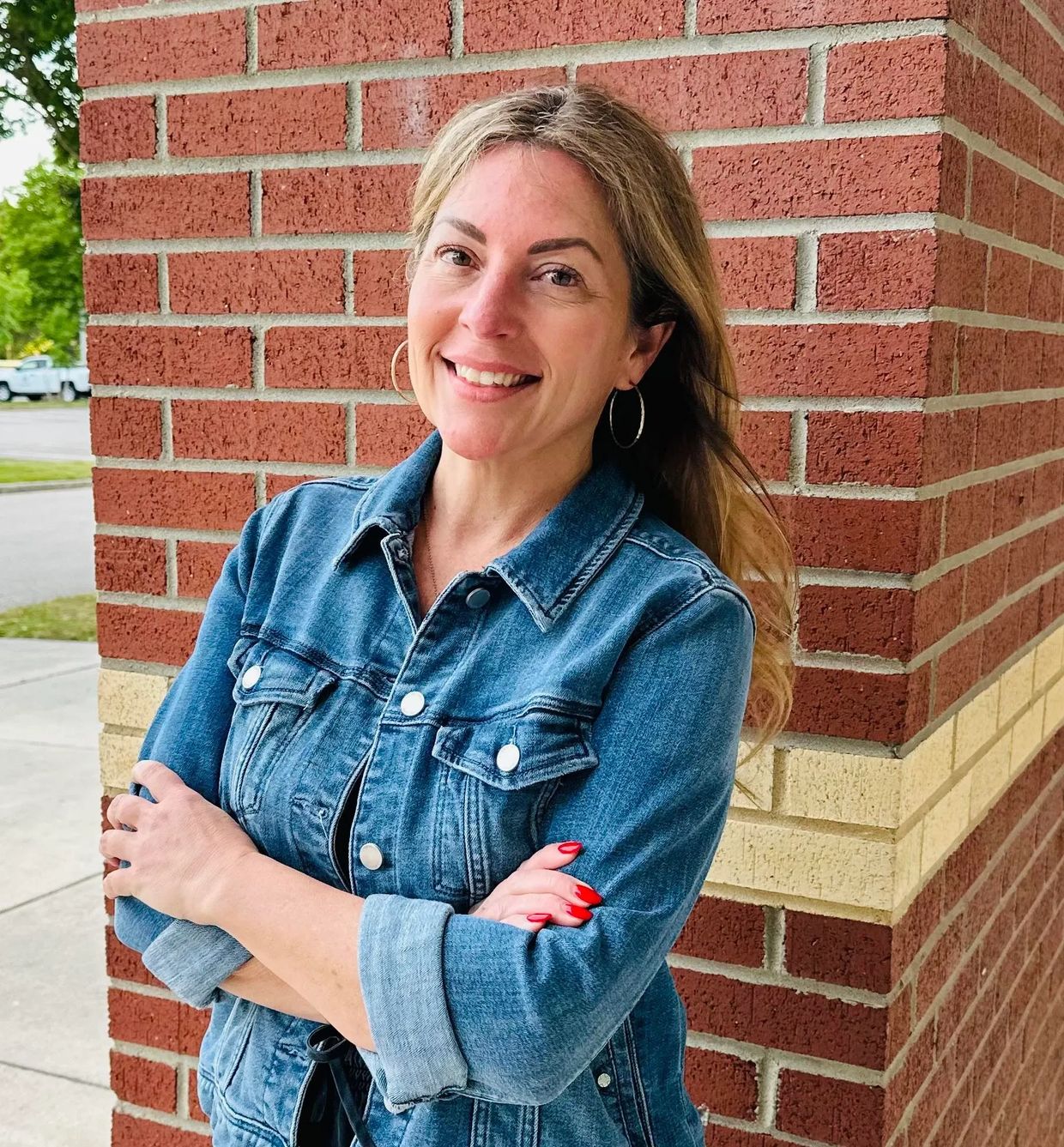 Smiling woman in a denim jacket standing against a brick wall.