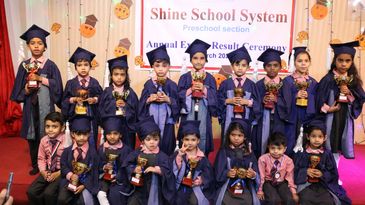Young students in graduation gowns holding trophies at a preschool awards ceremony.