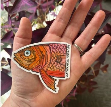 Hand holding a colorful fish-shaped sticker with artbyaleslud.com text.
