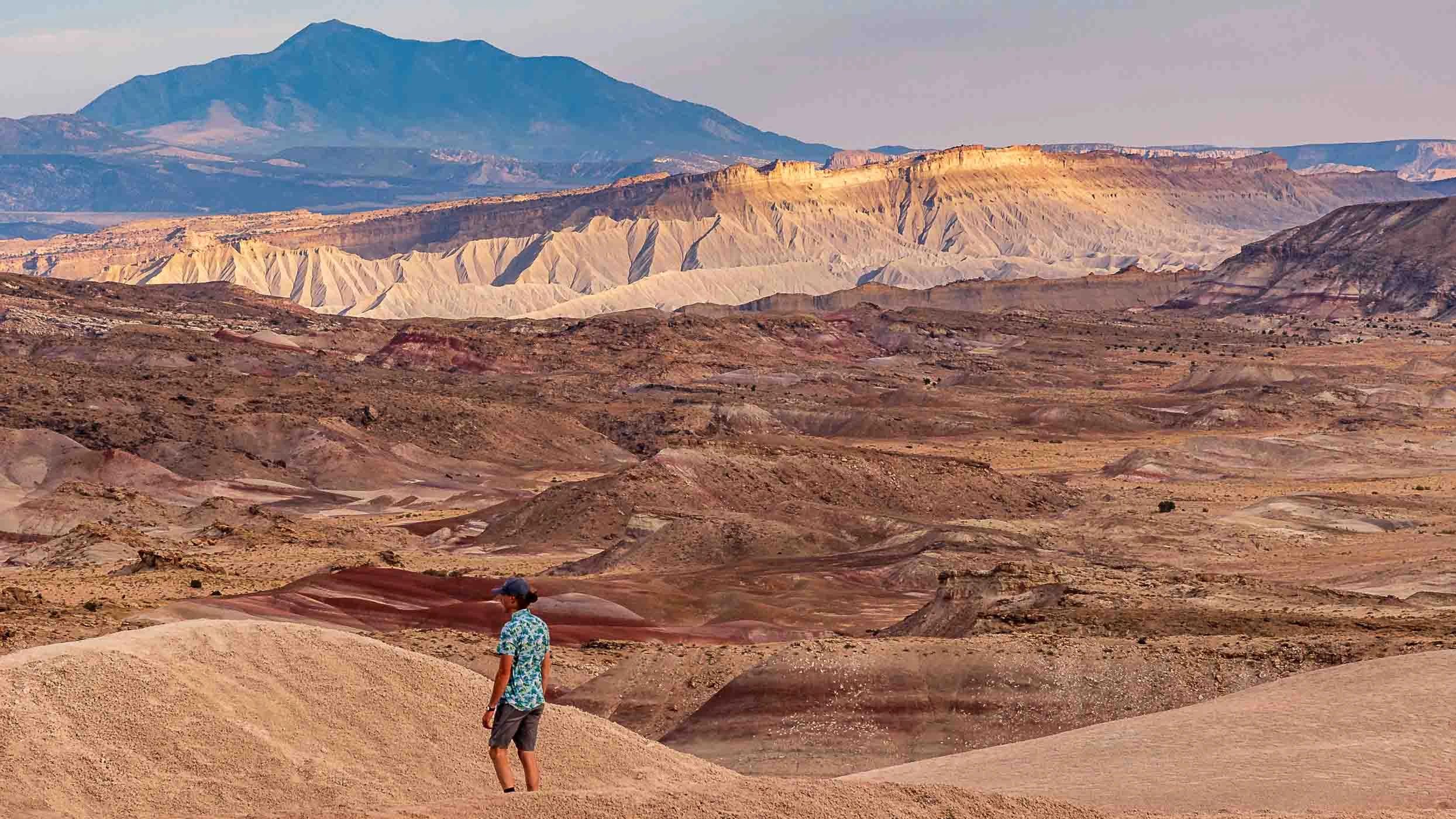 Person hiking in a colorful desert landscape with layered hills and distant blue mountains.