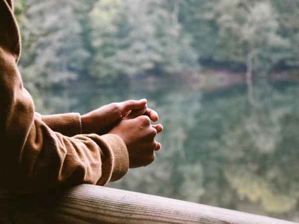 Person with clasped hands leaning on a wooden railing overlooking a calm lake.