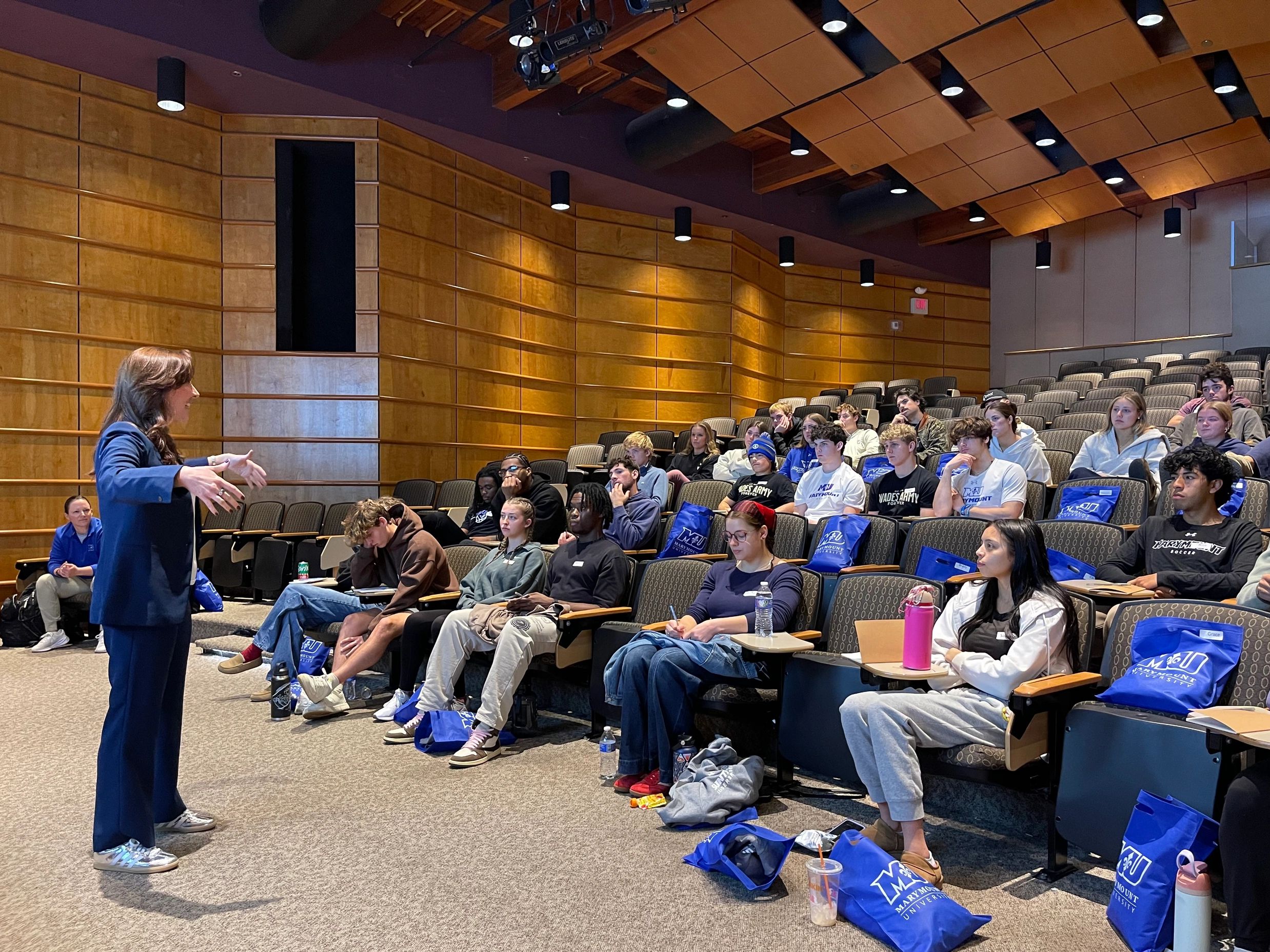 A woman speaks to attentive students in a modern lecture hall.