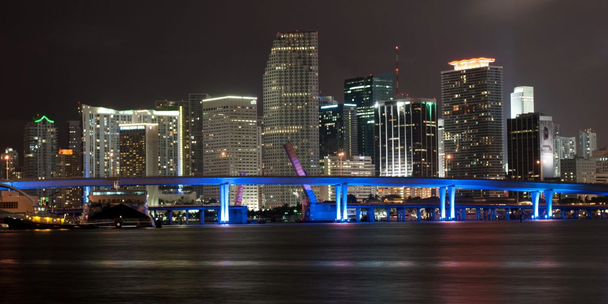 Night view of a city skyline with illuminated buildings and a blue-lit bridge over water.