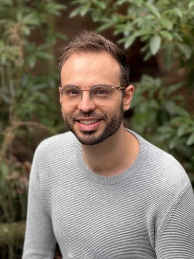 Smiling man in glasses and gray sweater outdoors with greenery background.