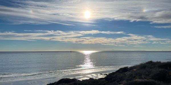 Sun shining over a calm ocean with a sandy beach and coastal vegetation.