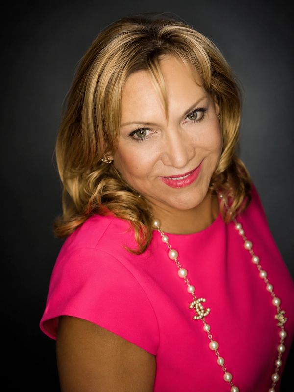 Smiling woman in a vibrant pink dress with pearl necklace against dark background.
