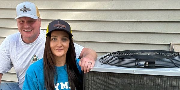A smiling couple posing by an outdoor air conditioning unit in casual clothing and hats.