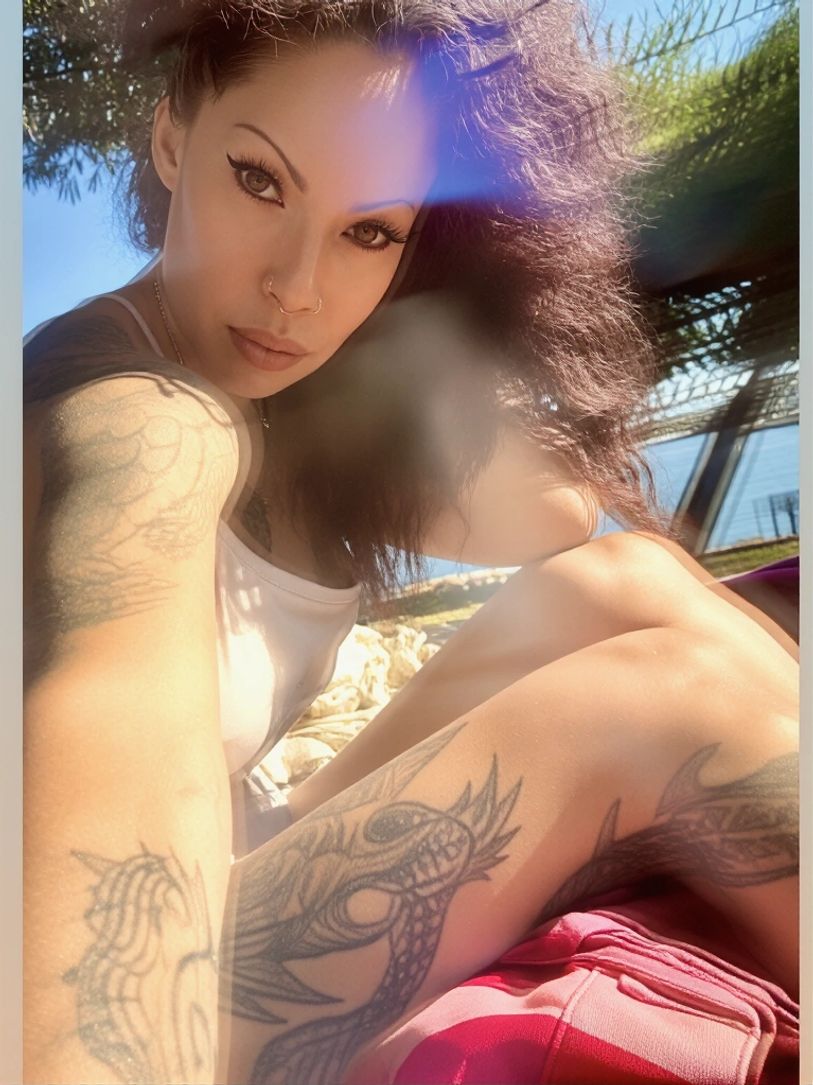Woman with tattoos poses outdoors in sunlight, with striking eye makeup and curly hair.