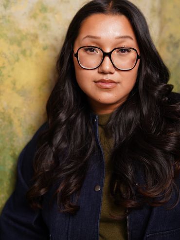 Young woman with long dark hair and glasses in front of a textured yellow-green wall.