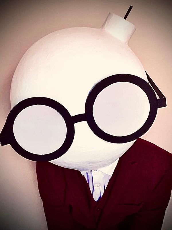 Person wearing a large round white mask with oversized black glasses and a burgundy suit.